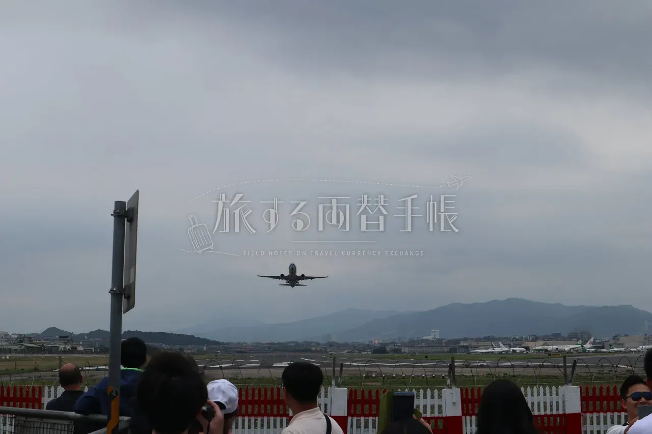 松山空港の離陸滑走路と背景の山並み。赤白柵越しに飛行機を見る人々