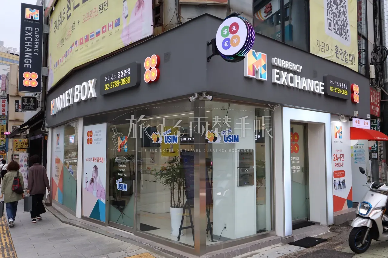 明洞 MoneyBox 本店の角地ファサード。丸いドット看板と 24시간 환전된다の広告